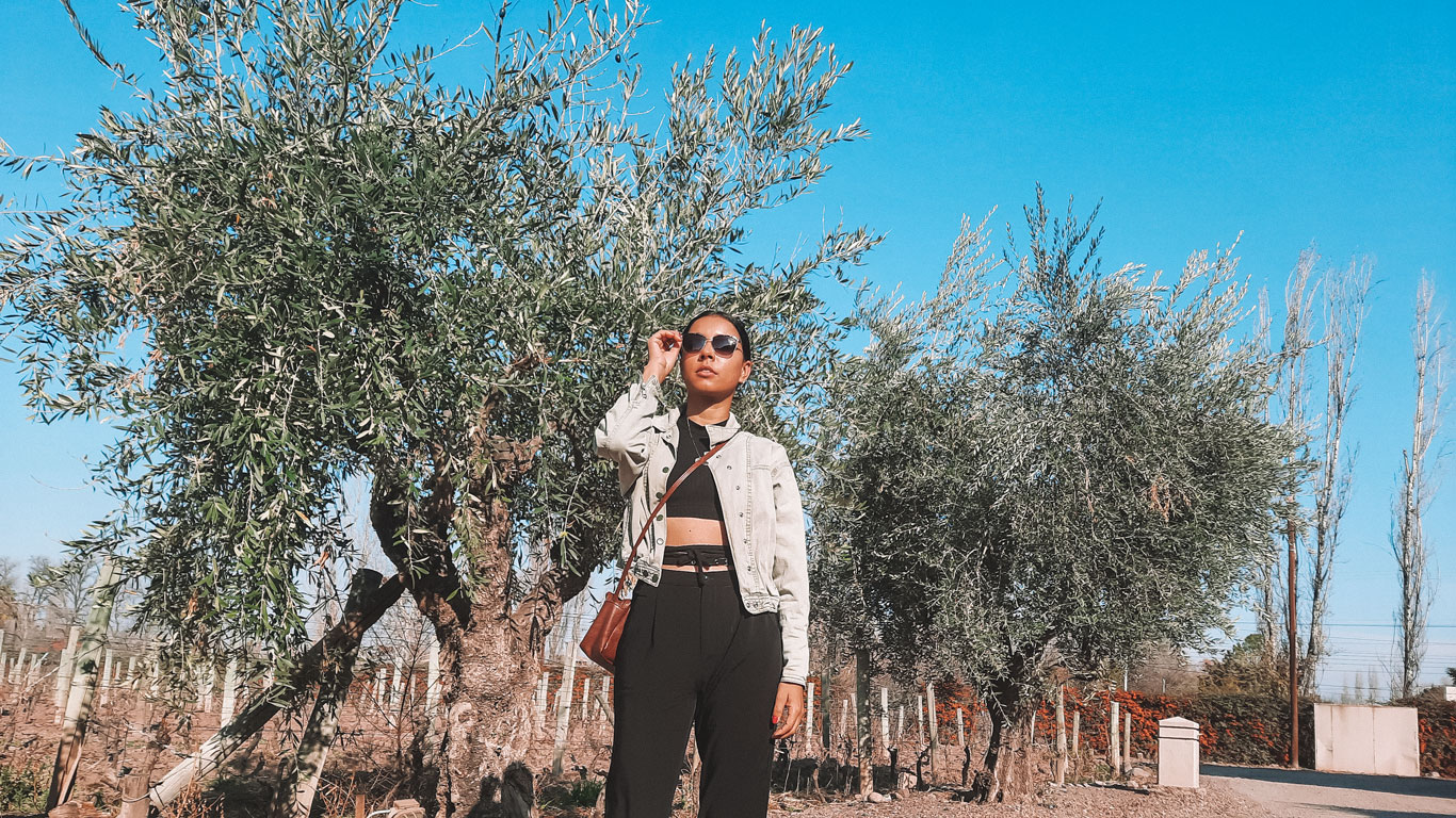 A person stands confidently in front of lush olive trees under a clear blue sky, wearing a stylish outfit consisting of a cropped black top, light denim jacket, and black pants. They adjust their sunglasses, and a brown crossbody bag hangs at their side. The setting appears to be a vineyard or orchard, suggesting a sunny day in a rural or agricultural region.