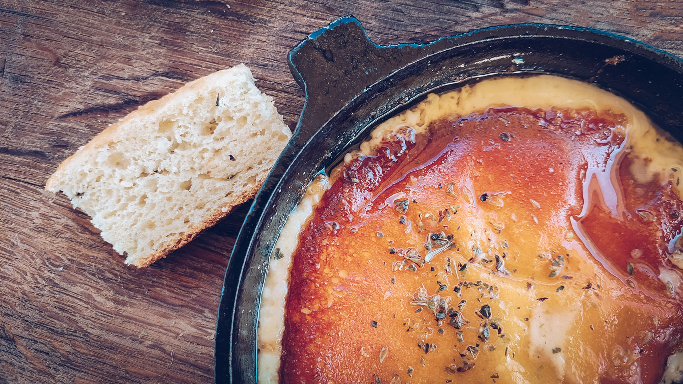 A close-up of a sizzling hot skillet filled with melted, golden-brown cheese topped with herbs and spices. A slice of crusty bread sits beside the skillet on a rustic wooden table, ready for dipping. The dish appears rich and savory, suggesting a classic comfort food or appetizer experience.