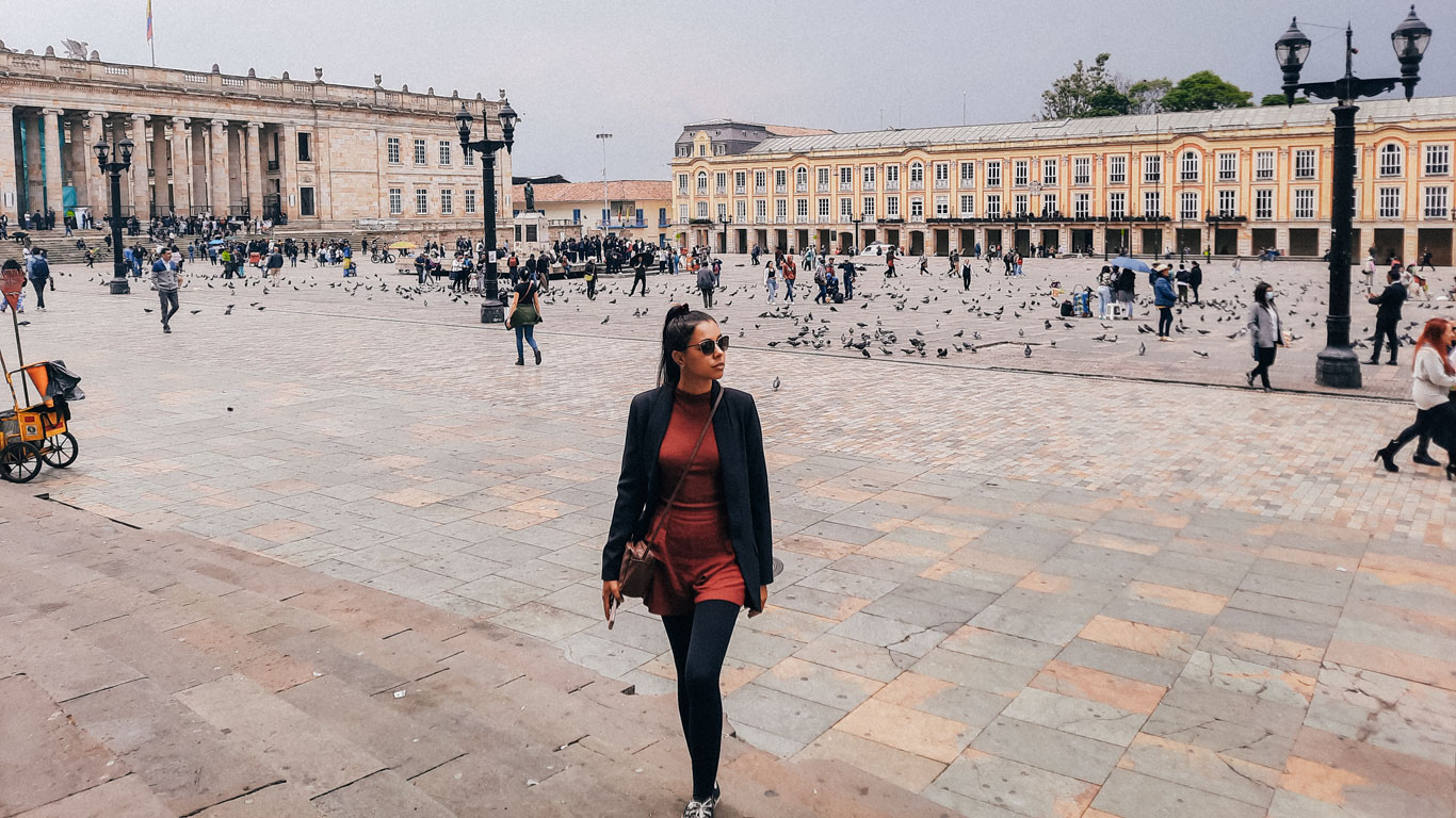 A woman in sunglasses and a black blazer walks across a spacious plaza paved with stone tiles, surrounded by pigeons and people. In the background, there's a historic yellow building with columns and multiple windows, with groups of people gathered near steps and statues. The scene is set in Bogotá, Colombia, capturing an urban setting filled with activity.