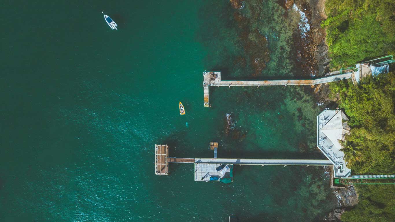 This aerial image shows a serene view of piers extending into the emerald-green waters of Salvador, Brazil. A couple of small boats float nearby, while lush greenery lines the coastline. The clear water reveals rocky formations beneath, creating a tranquil and picturesque scene.