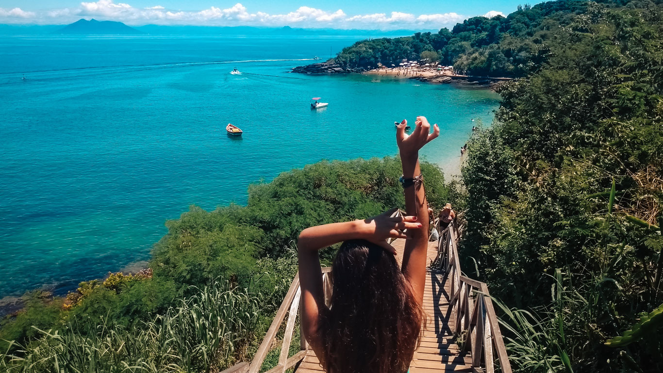 This image captures a scenic descent to Azedinha Beach, with a woman standing on a wooden staircase, stretching her arms as she takes in the view. The turquoise waters below are dotted with small boats, surrounded by lush greenery and distant mountains under a bright blue sky. 