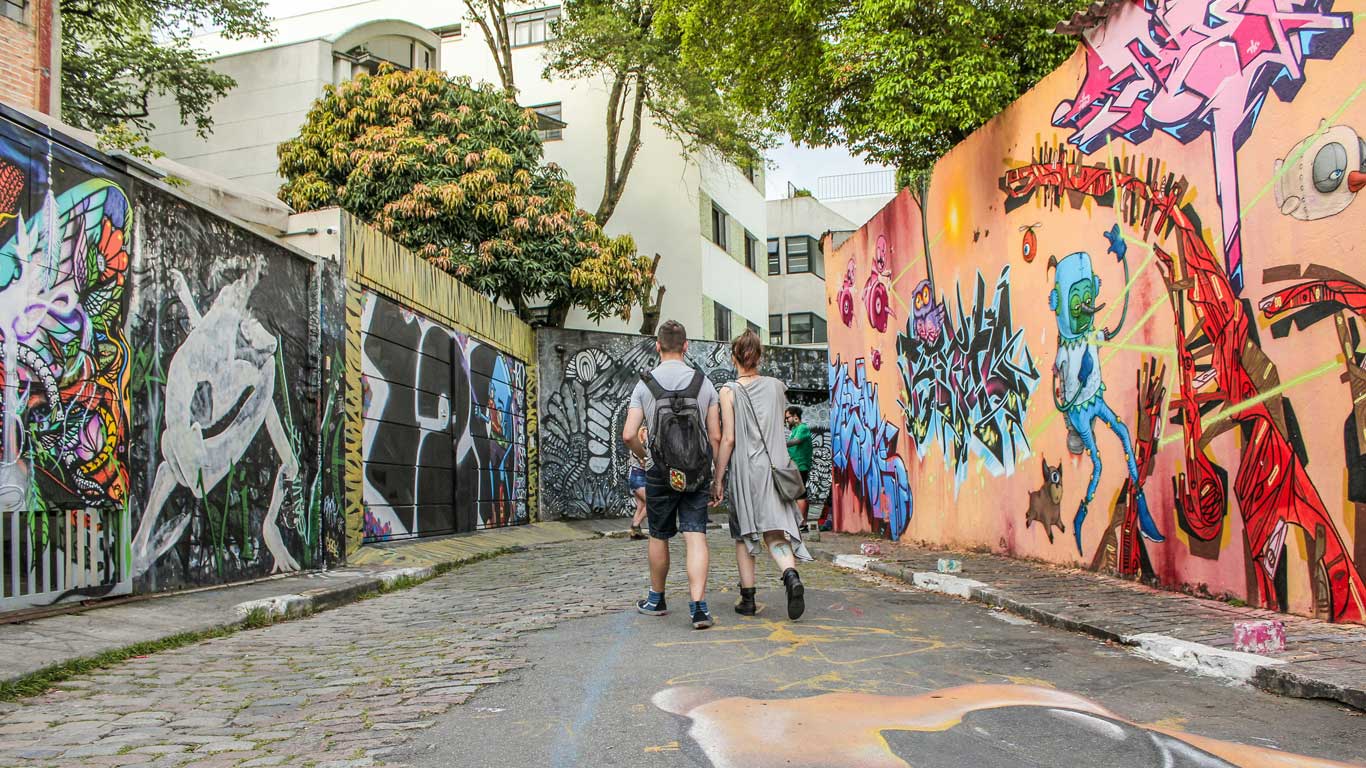 This image depicts Beco do Batman, a vibrant alley in São Paulo adorned with colorful and eclectic street art murals on both sides. Two people are seen walking along the cobblestone path, surrounded by bold graffiti designs, showcasing the artistic and creative atmosphere of this iconic urban destination.