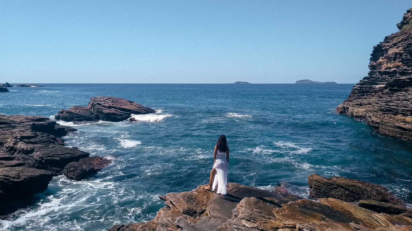 This image features a woman standing on rocky cliffs overlooking a vast, calm ocean with gentle waves crashing against the rocks. The scene is serene, with clear blue skies and small distant islands visible on the horizon.