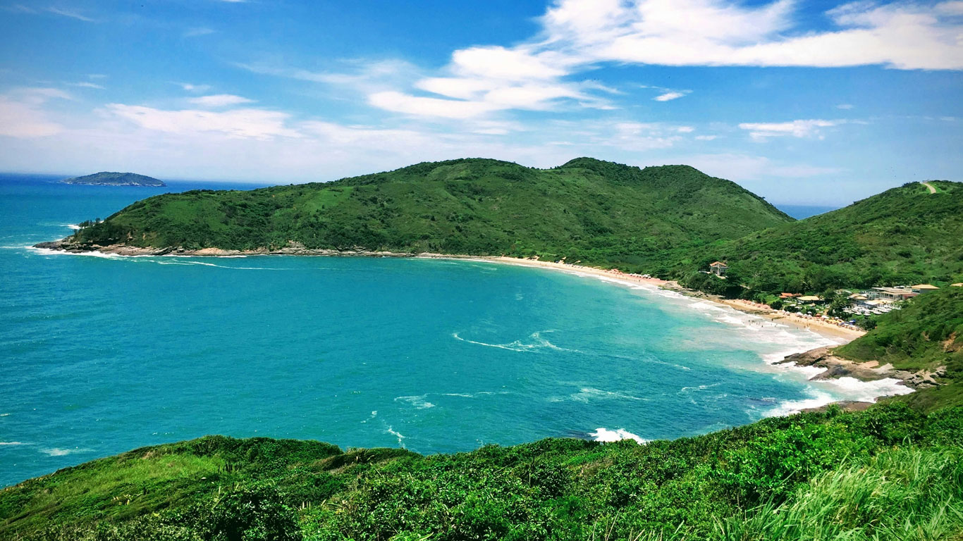 This image showcases Brava Beach, nestled in a curved bay surrounded by lush green hills and turquoise waters. The sandy beach stretches along the shoreline with small waves gently breaking, while a few scattered buildings and beachgoers are visible near the right side. 