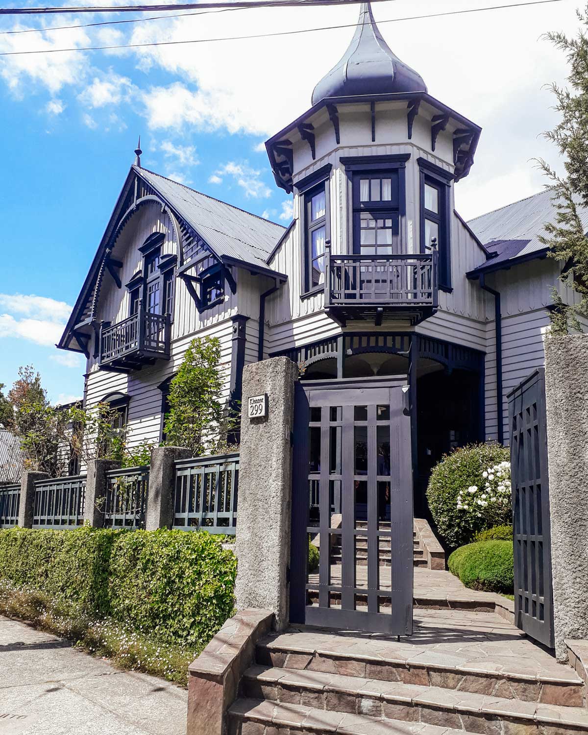 This image features a charming, historic-style house in Puerto Varas, Chile, with a turret, steep gabled roofs, and detailed wooden accents. The exterior is painted in a light color with dark trim, and a small garden and paved pathway lead to the open gate. It reflects the region's distinctive architecture and welcoming atmosphere.