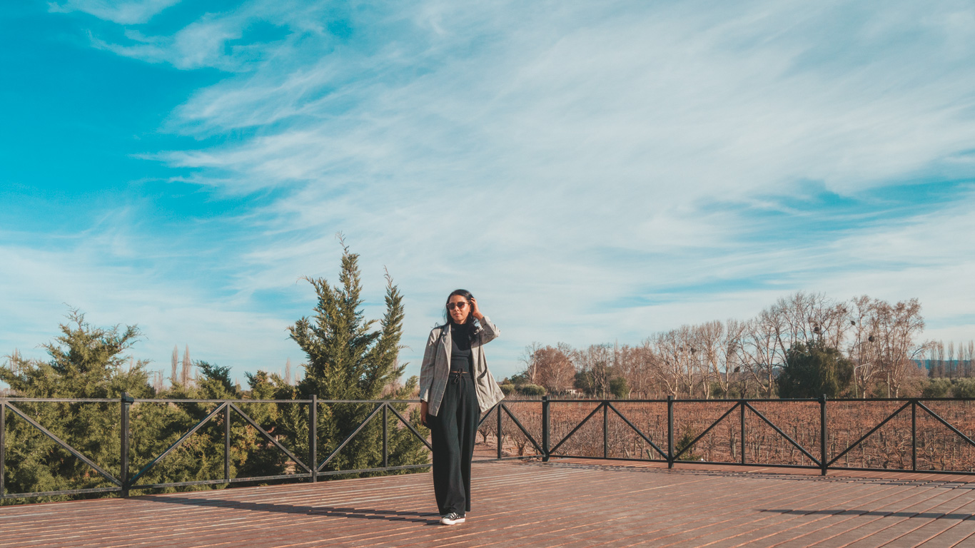 A woman standing on a wooden deck with a backdrop of green trees and bare vineyards under a vivid blue sky. She is dressed in a blazer, dark pants, and sunglasses, exuding a casual yet chic style. The serene setting highlights the open-air charm of a vineyard landscape.