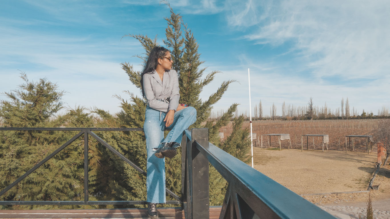 A woman sitting on a black metal railing, wearing a gray blouse, blue jeans, and sunglasses, with a scenic backdrop of vineyards and tall green trees under a bright blue sky. The scene exudes tranquility, with wooden trellises and a vineyard landscape stretching into the distance.