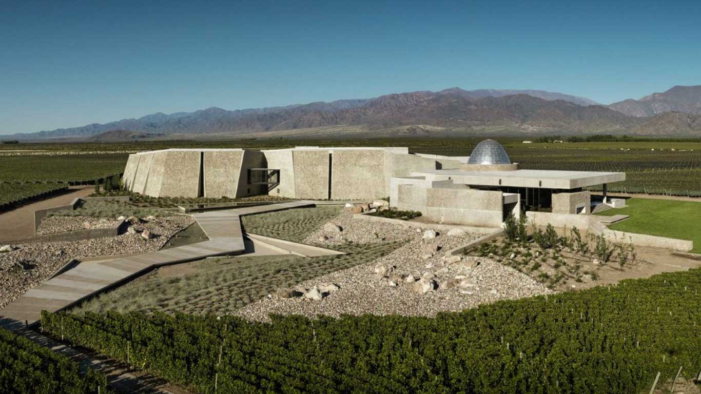 The modern and minimalist architecture of Bodega Zuccardi in Mendoza, Argentina, featuring circular stone structures and a domed roof. The winery is surrounded by expansive vineyards with the Andes mountains providing a striking backdrop under a clear blue sky. The scene highlights the harmony between contemporary design and the natural landscape.