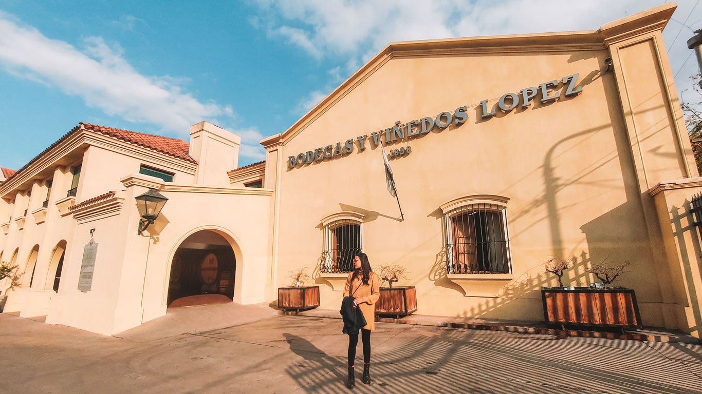 A woman standing in front of the Bodegas y Viñedos López building, a cream-colored winery with arched windows and a prominent sign displaying its name and founding year, 1898. She is wearing a tan coat and black leggings while holding a jacket, with a clear blue sky adding to the warm, inviting atmosphere.