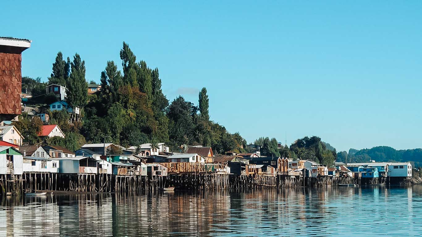 This image captures the iconic "palafitos" in Castro, Chiloé—colorful wooden houses built on stilts along the waterfront. The calm water reflects the homes and surrounding lush greenery, creating a serene and picturesque scene. 