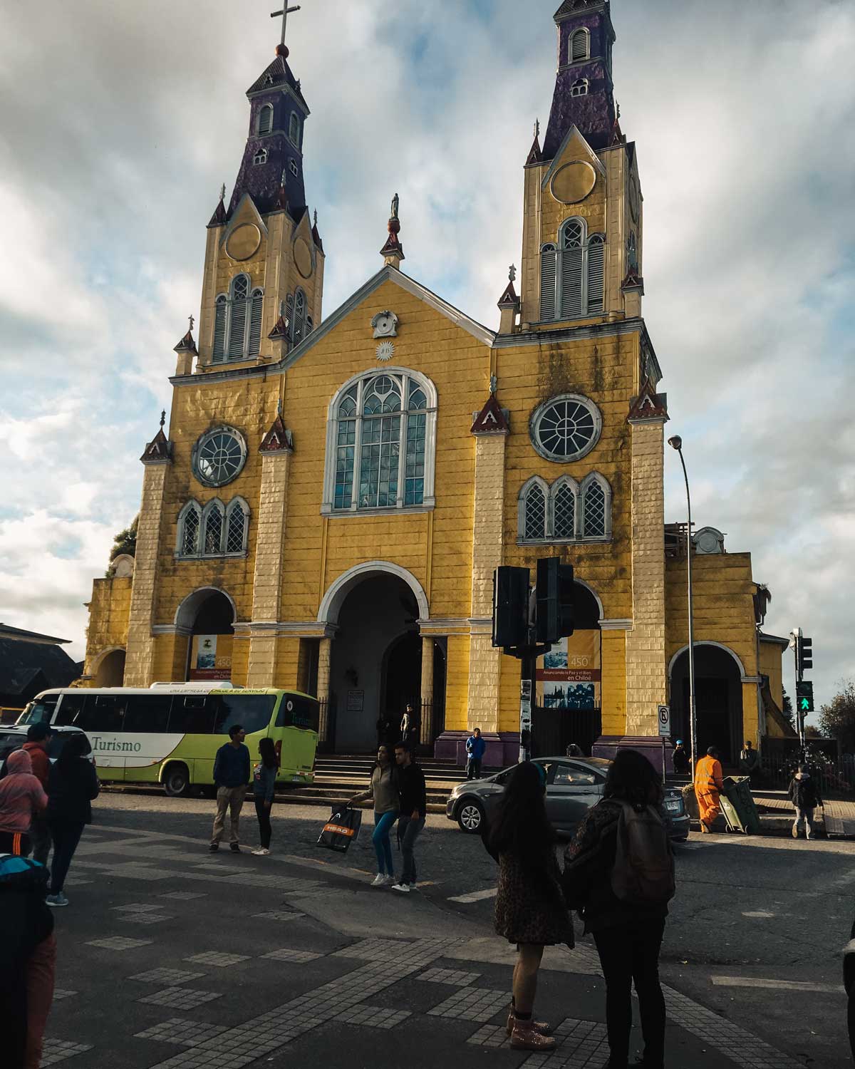 This image showcases the colorful San Francisco Church in Castro, Chiloé, with its striking yellow façade and twin purple spires. The church's detailed stained-glass windows and architectural beauty are complemented by a busy street scene with tourists and locals. 