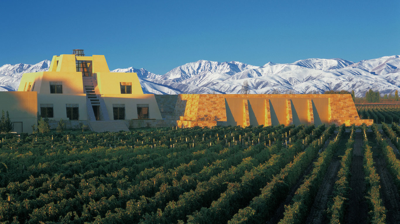 The iconic Catena Zapata winery in Mendoza, Argentina, designed like a Mayan pyramid, surrounded by lush green vineyards. Behind the winery, the Andes mountains stand majestically, covered in snow, contrasting against the deep blue sky. The scene captures the elegance and grandeur of this renowned winery.