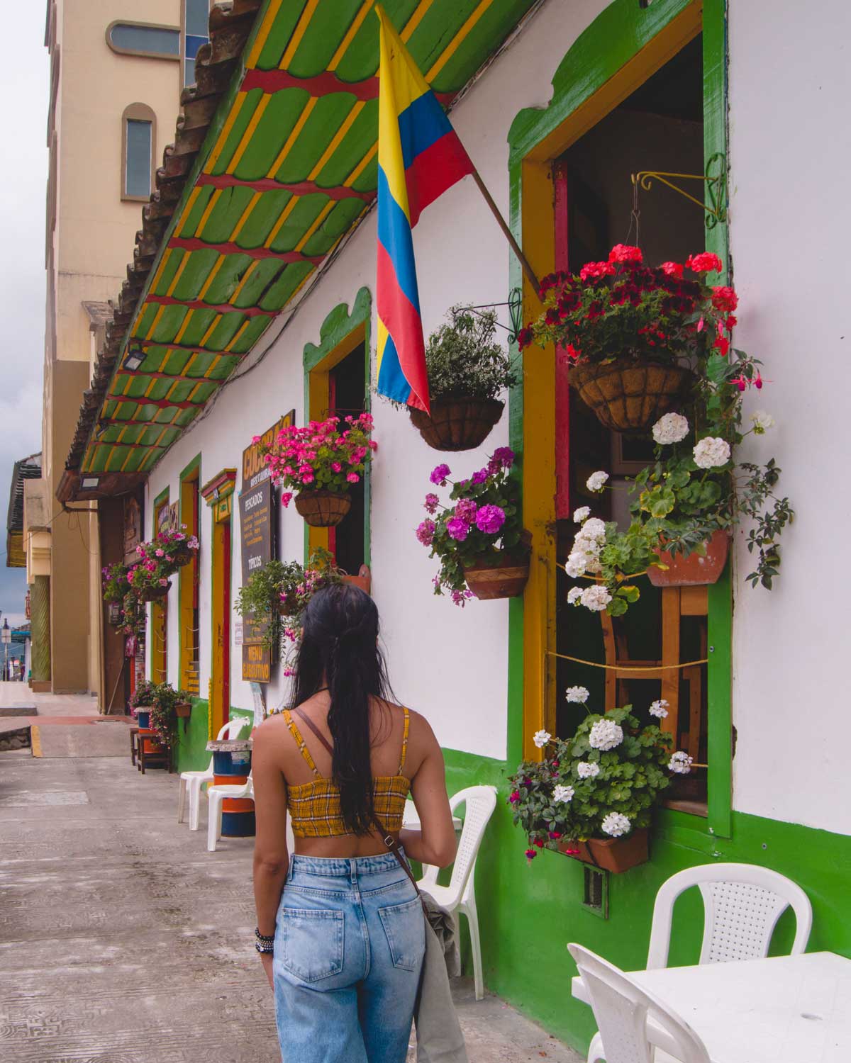 This image captures a woman with long black hair wearing a yellow plaid crop top and jeans walking past a colorful building in Salento. The building features vibrant green and yellow accents, hanging flower baskets bursting with bright blooms, and a Colombian flag proudly displayed, showcasing the town's charm and cultural pride.