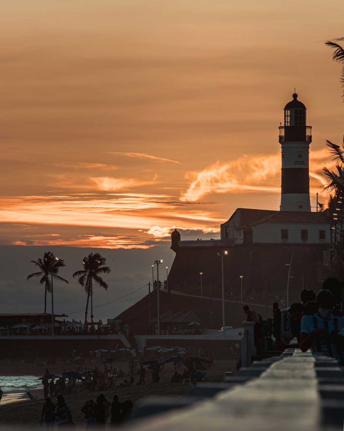 This image captures the Farol da Barra lighthouse in Salvador, Brazil, silhouetted against a dramatic orange sunset. The scene includes a lively beach with people enjoying the evening, framed by palm trees and warm light reflecting on the horizon.