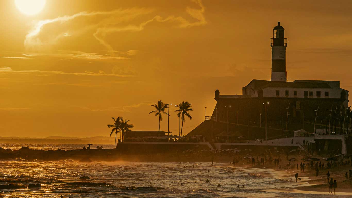 This image captures the Farol da Barra lighthouse in Salvador, Brazil, silhouetted against a dramatic orange sunset. The scene includes a lively beach with people enjoying the evening, framed by palm trees and warm light reflecting on the horizon.