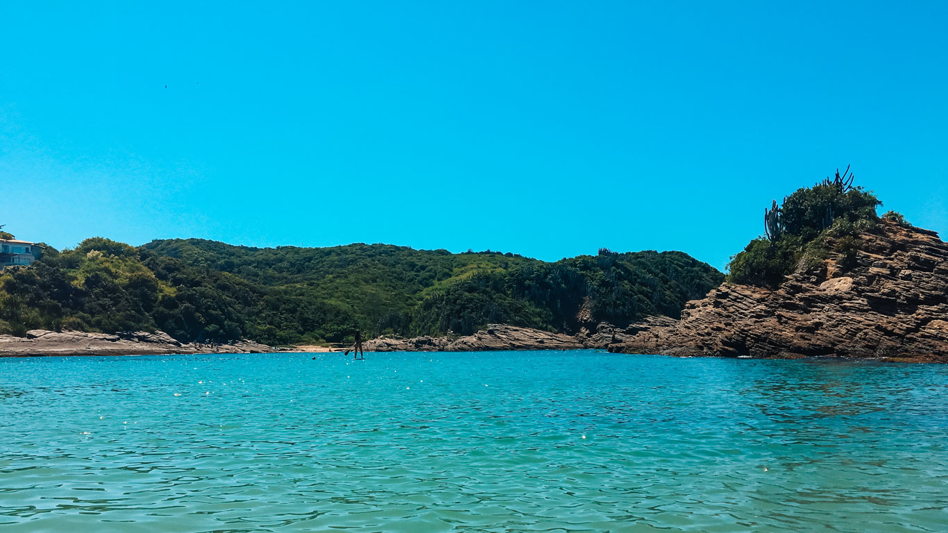 This image shows a tranquil beach with crystal-clear turquoise water and lush green hills in the background. A paddleboarder glides across the calm surface, and rocky outcroppings frame the scene, adding to the tropical and serene ambiance.