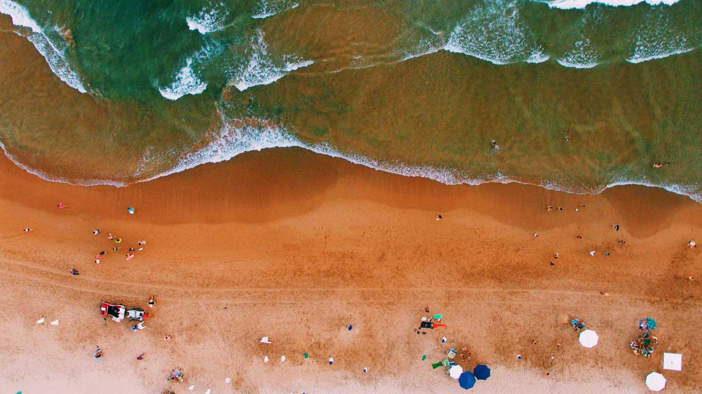 This aerial view of Forno Beach highlights its vibrant orange sand and the emerald waves lapping the shore. Beachgoers are scattered across the sand, some under umbrellas and others enjoying the water, creating a dynamic and colorful scene.