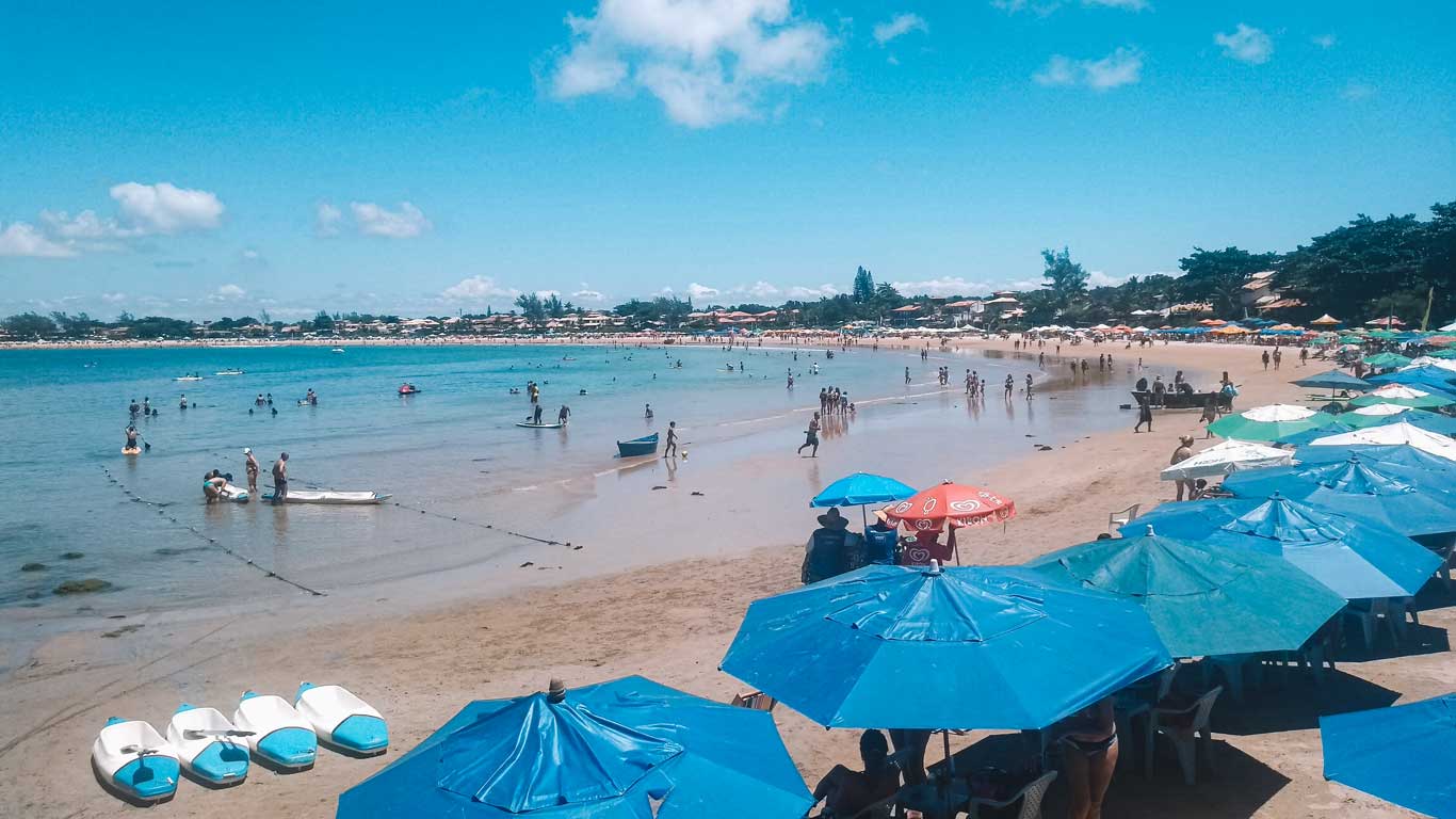 This image shows Geribá Beach bustling with activity, featuring a wide sandy shoreline dotted with colorful umbrellas and beachgoers enjoying the sun. The calm, shallow water is filled with swimmers, paddleboarders, and small boats, while houses and greenery line the distant shore under a bright blue sky.