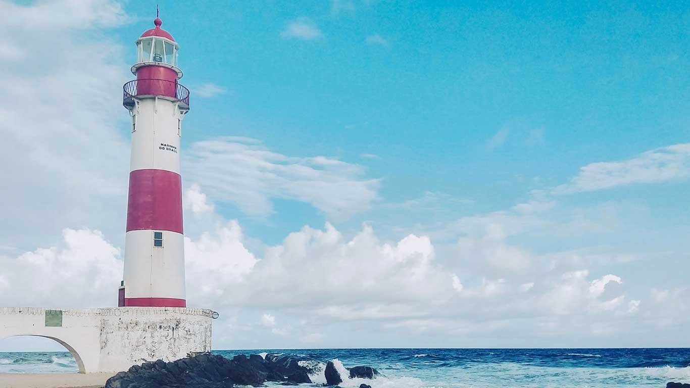 This image captures the Itapoã Lighthouse in Salvador, Brazil, with its red and white stripes standing tall against a bright blue sky. Waves crash onto the rocks at its base, creating a dynamic contrast to the serene horizon of the ocean. The lighthouse is a picturesque symbol of coastal beauty and navigation.