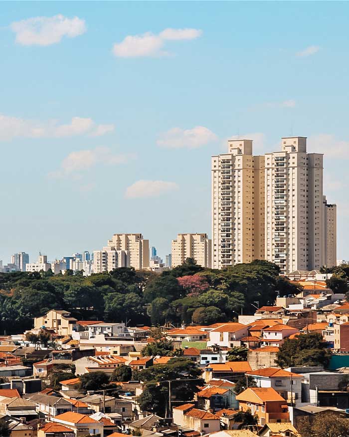 This image features a view of São Paulo, showcasing a mix of residential areas with red-tiled roofs in the foreground and modern high-rise apartment buildings in the background. The cityscape is complemented by patches of green trees under a bright blue sky with scattered clouds, reflecting the blend of urban and natural elements in the city.