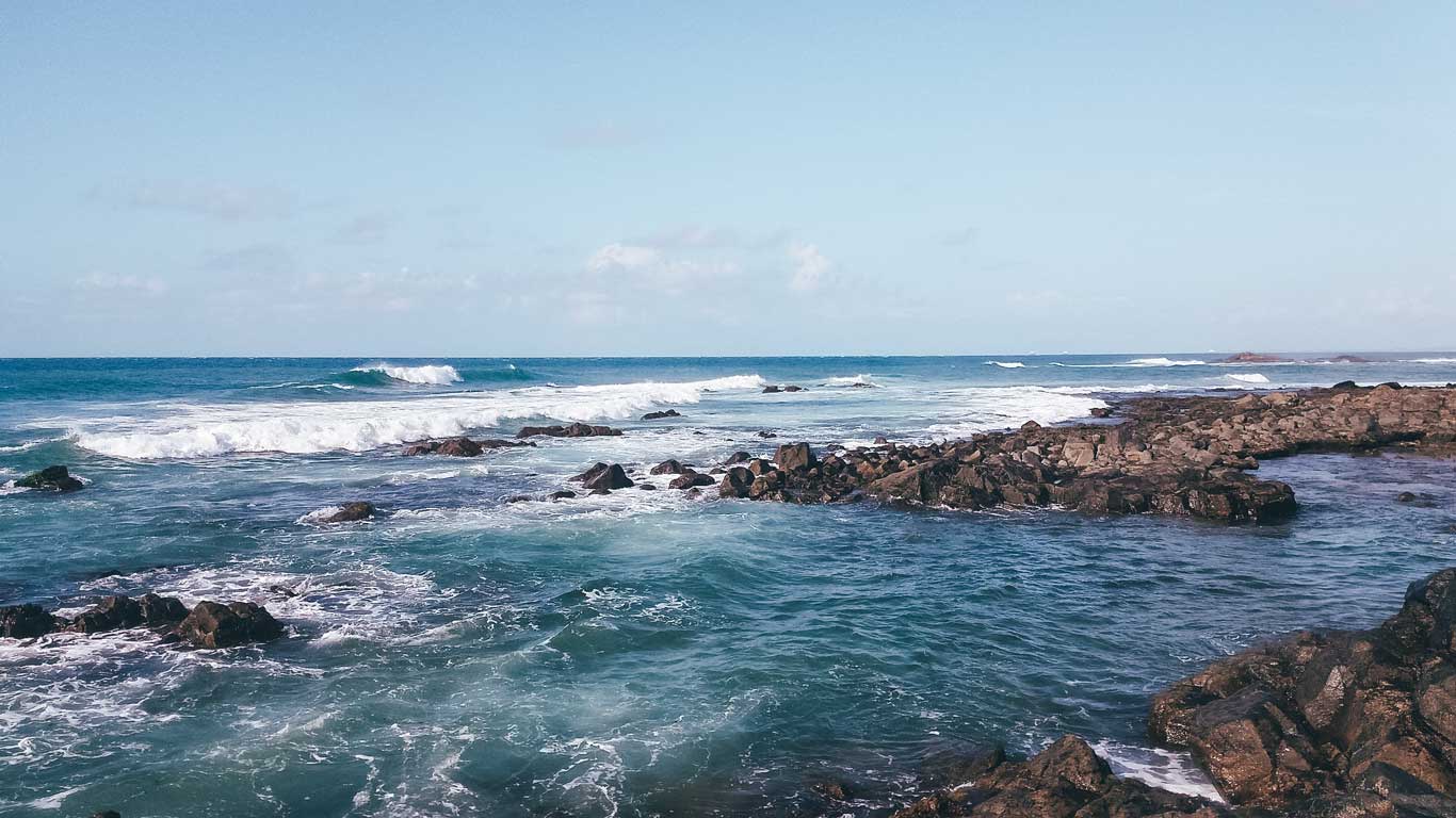 This image shows a rocky coastline in the Ondina neighborhood with gentle waves crashing against the shore under a clear blue sky, an excellent choice for where to stay in Salvador. The turquoise waters sparkle in the sunlight, creating a tranquil and scenic view of the ocean.