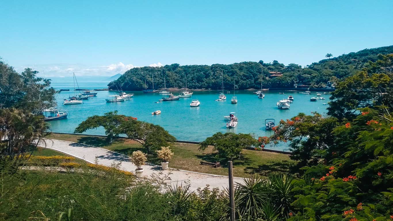 This image captures Ossos Beach, a charming bay dotted with anchored boats and surrounded by lush green hills. A well-maintained garden with vibrant trees and pathways frames the foreground, adding a serene and picturesque vibe. 