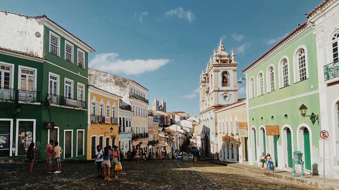 This image showcases the vibrant streets of Pelourinho, Salvador's historic center, lined with colorful colonial buildings in shades of green, yellow, and blue. The cobblestone road leads to the ornate façade of a baroque-style church, while groups of people enjoy the lively and cultural atmosphere. The scene highlights the area's rich history and charm under a bright blue sky.