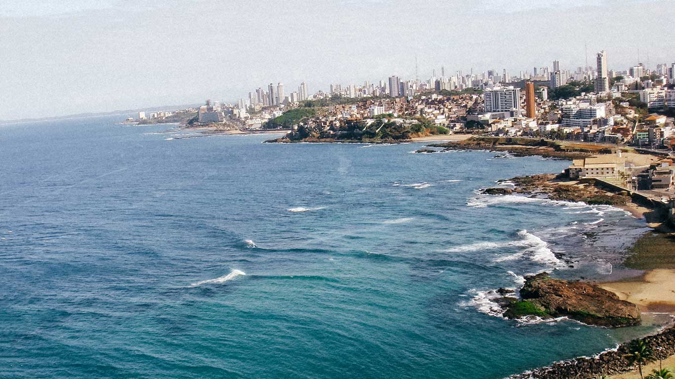 This image showcases the coastline of Salvador, Brazil, with its turquoise waters meeting the rocky shores and sandy beaches. The cityscape in the background features a mix of modern high-rises and traditional architecture, with lush greenery dotting the urban landscape. The view highlights the vibrant charm of the Rio Vermelho district along the ocean.