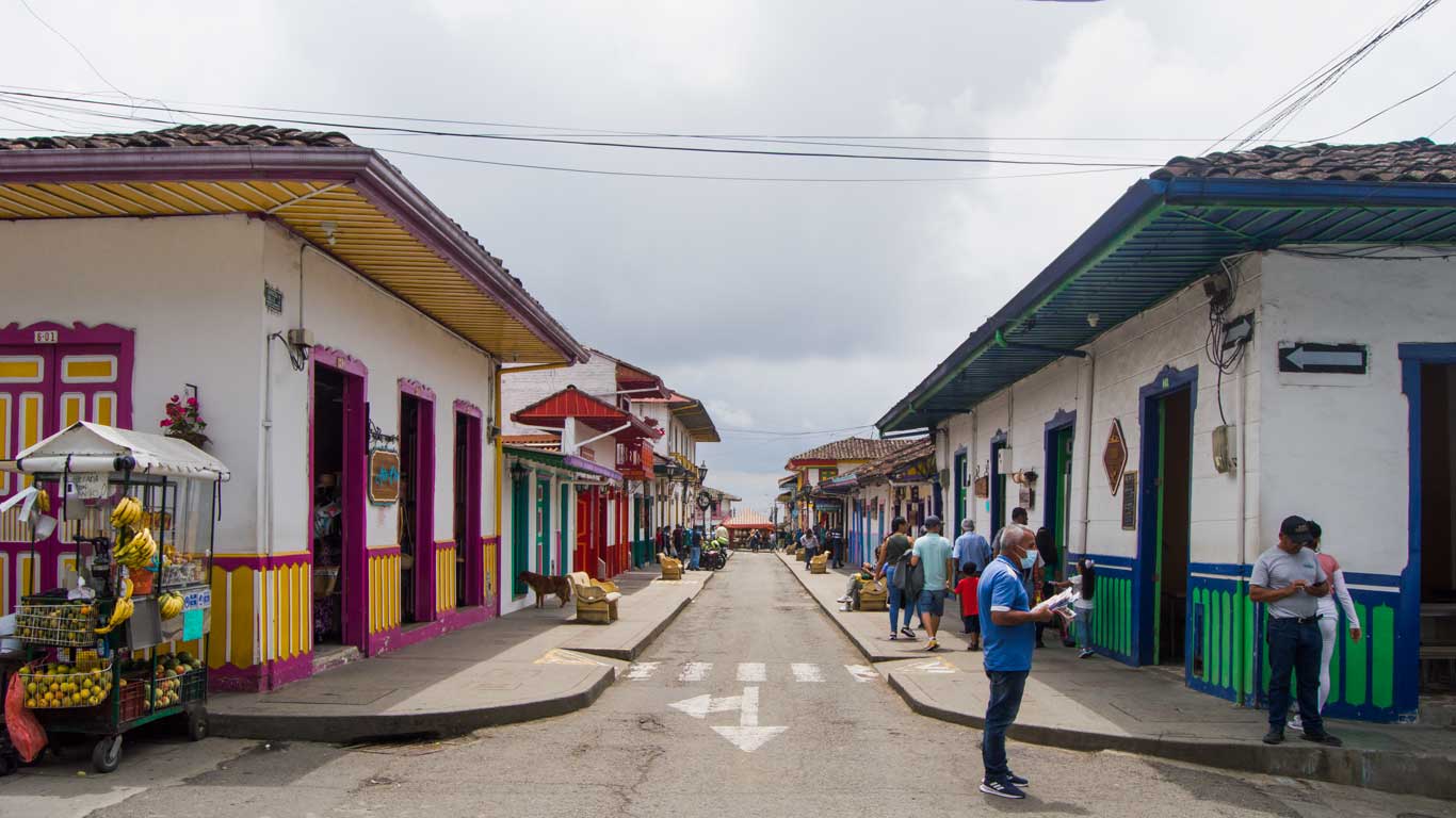 This image shows a colorful street in Salento, lined with traditional buildings painted in vibrant shades of white, pink, blue, and yellow, with decorative wooden accents. A street vendor's cart with fruits like bananas and oranges is visible on the left, while people stroll and interact along the street, capturing the lively and charming atmosphere of the town.