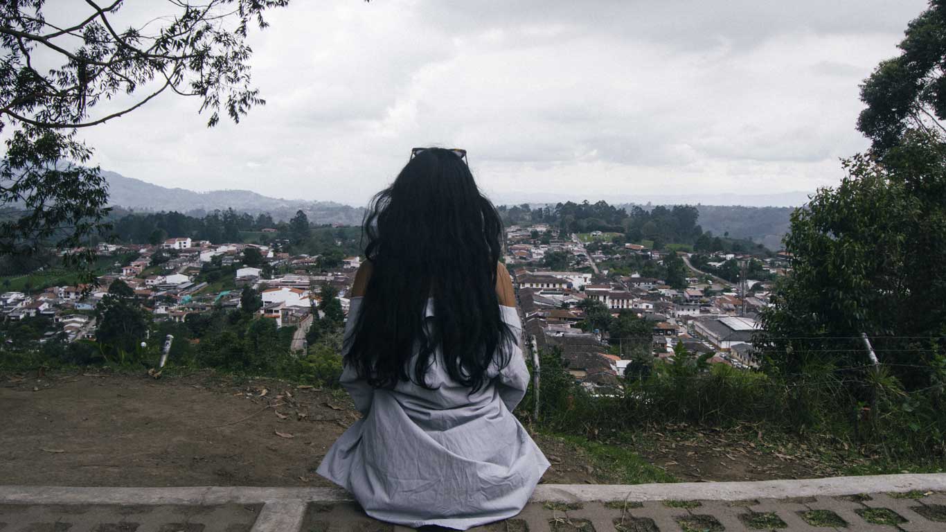 This image shows a person with long black hair, wearing a light-colored shirt, sitting on a ledge and looking out over Salento Village nestled among green hills under a cloudy sky. The scene conveys a serene and contemplative moment, with a picturesque view of the town surrounded by nature.