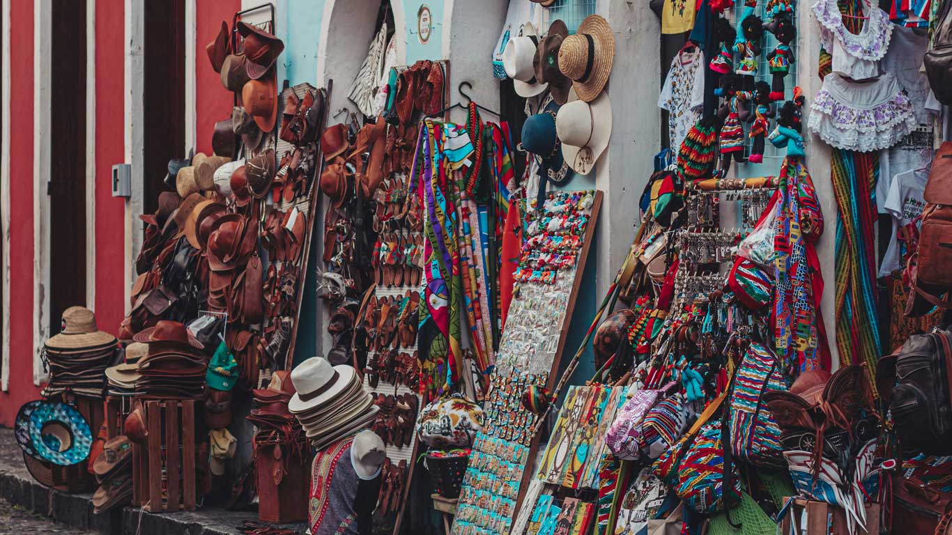 This image depicts a colorful street market in Salvador, Brazil, showcasing an array of handmade goods. The stalls are filled with hats, leather goods, colorful scarves, keychains, and traditional crafts, creating a vibrant and lively atmosphere. The items reflect the rich cultural heritage and artisanal craftsmanship of the region.