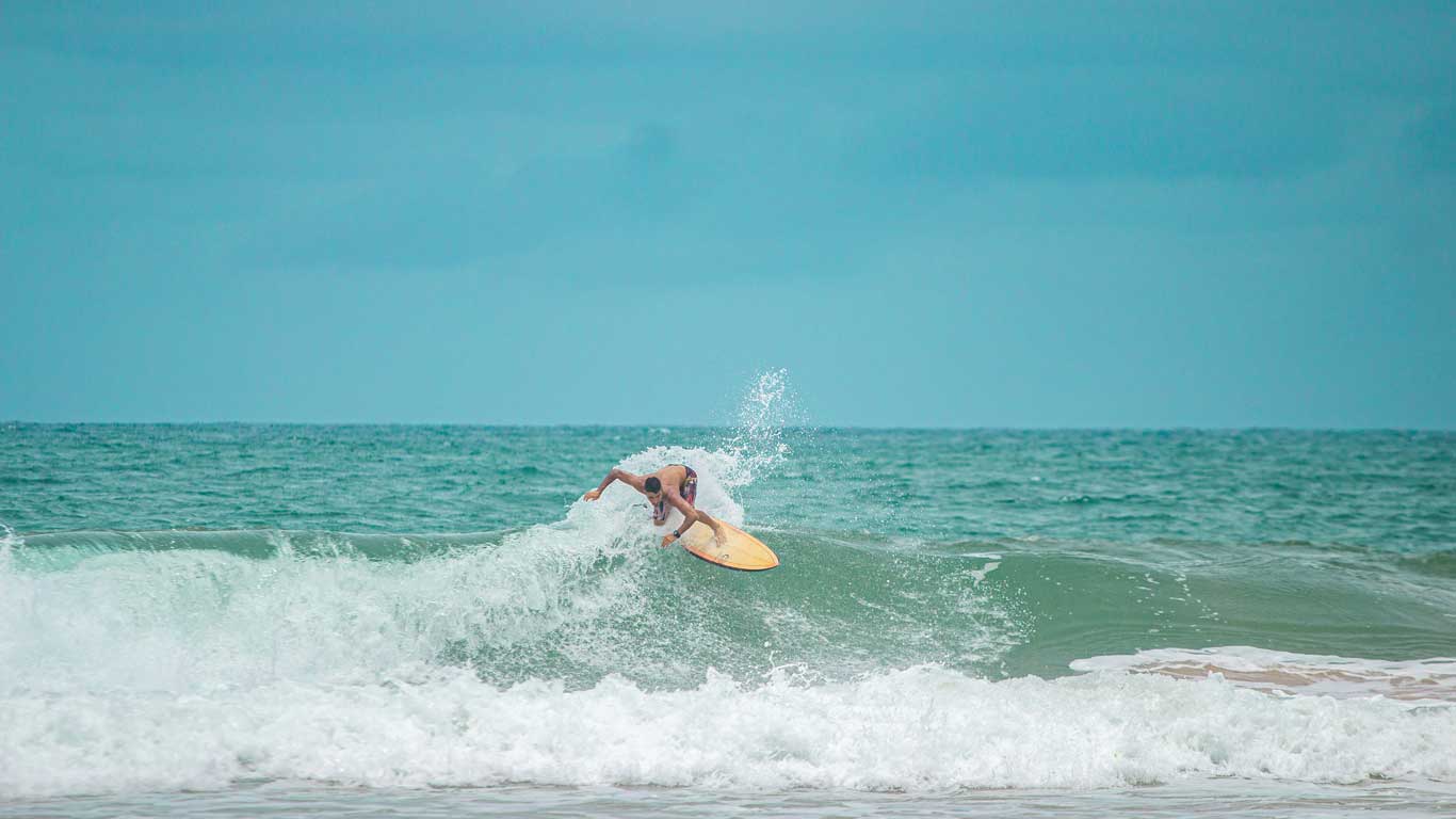 This image features a surfer riding a wave at Stella Maris Beach in Salvador, Brazil. The surfer leans into the crest of the turquoise wave, creating a dynamic splash against the calm ocean backdrop. The scene reflects the vibrant surf culture and natural beauty of the area.