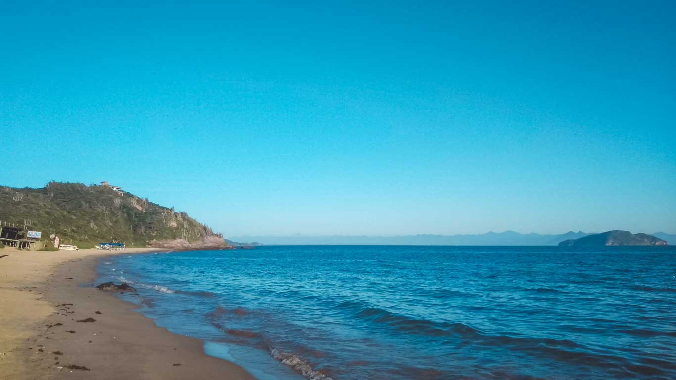 This image shows the serene Tartaruga Beach, featuring a quiet sandy shoreline that gently curves along the edge of calm blue waters. A rocky hill covered in greenery rises on the left, while distant islands and mountains are visible on the horizon under a clear blue sky.