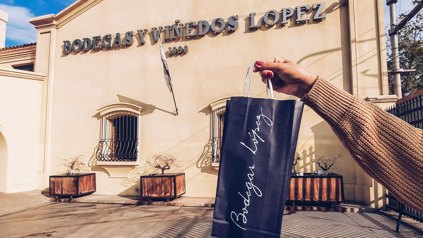 A hand holding a black shopping bag with the logo "Bodegas López" in front of the Bodegas y Viñedos López building. The cream-colored facade and the sign displaying the winery's name and founding year, 1898, are visible in the background. The image highlights a wine purchase and the elegant branding of the winery.