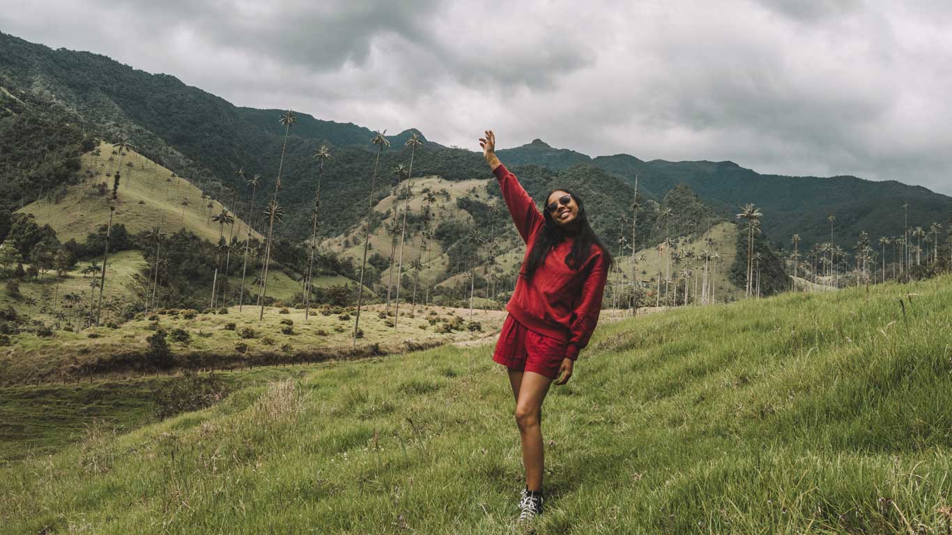 This image shows a woman dressed in a red outfit, smiling and posing with one arm raised in a lush green valley surrounded by rolling hills and towering wax palm trees, characteristic of the Cocora Valley near Salento. The dramatic cloudy sky and the serene natural landscape create a stunning backdrop for the scene.