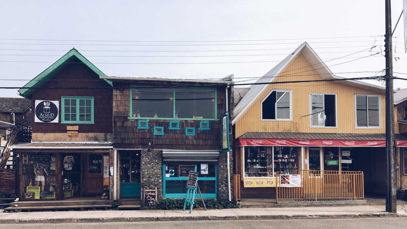 This image features a street view of two adjacent buildings with unique architecture in Chiloé. The left building has wooden shingles, bright teal window frames, and signage for "Almud," a local establishment, while the right building is painted yellow with a Coca-Cola-branded awning. Both structures house small businesses, blending rustic charm with a cozy commercial vibe.