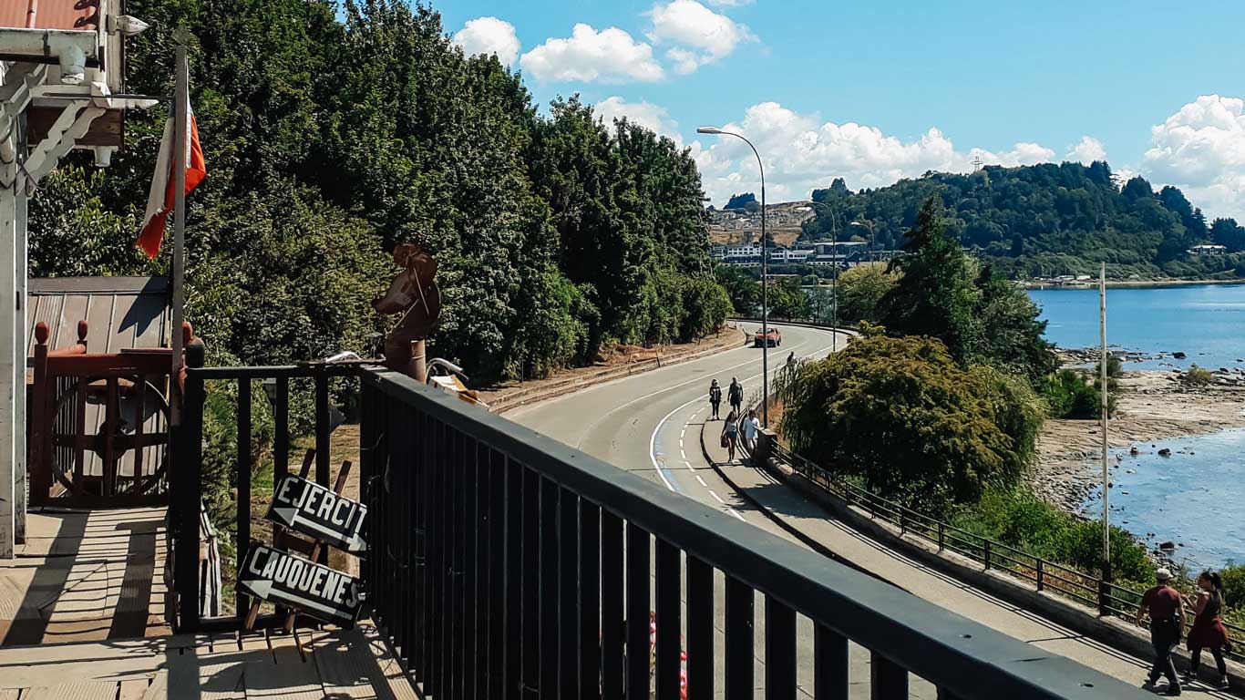 This image shows a scenic view of a waterfront road in Puerto Varas, Chile. In the foreground, there’s a wooden railing with signs and a Chilean flag, while the background features cyclists and pedestrians along a curved path surrounded by lush greenery and a calm lake. The image captures the charm of this vibrant and picturesque destination.