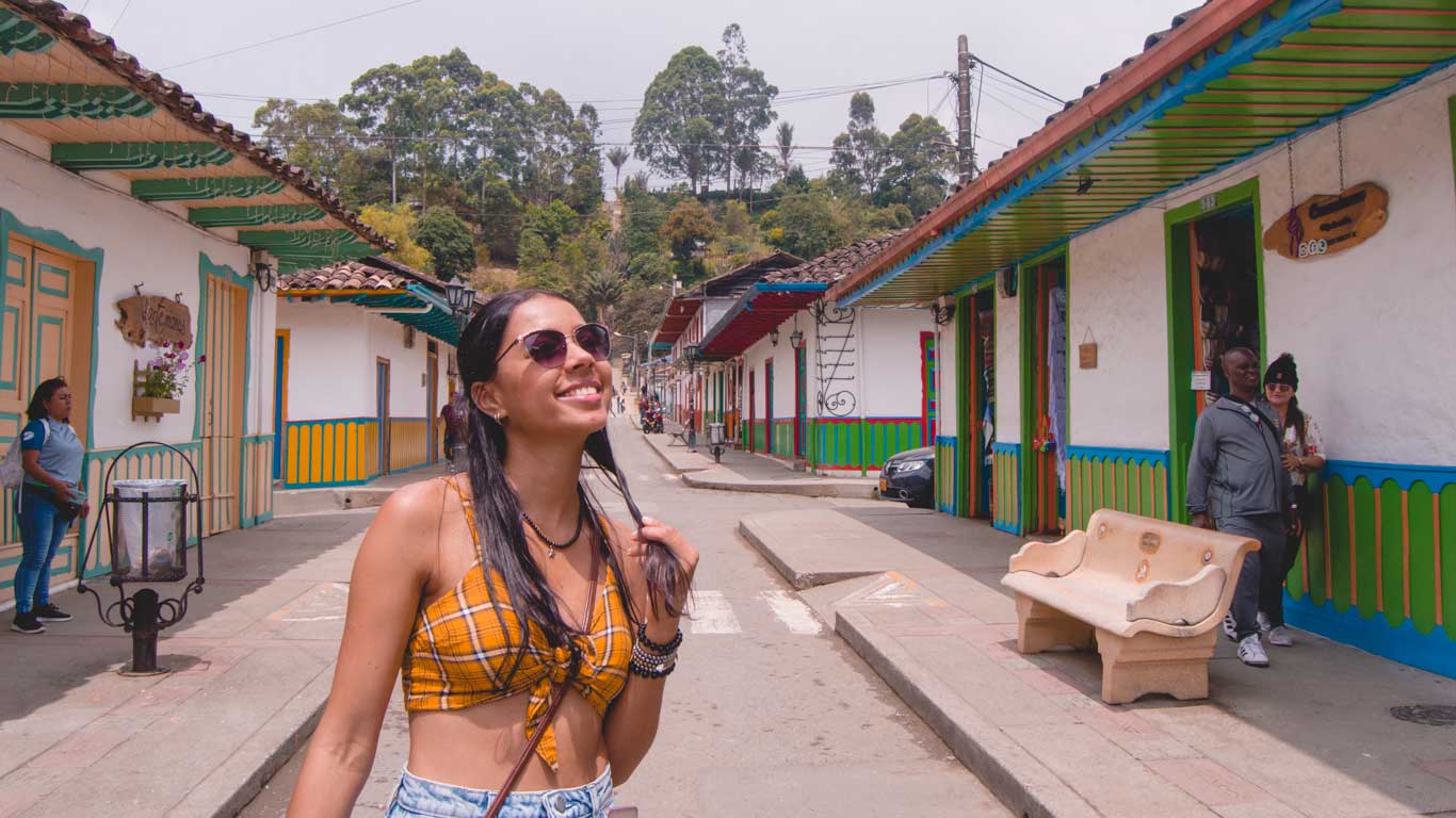 This image shows a smiling woman wearing sunglasses, a yellow plaid crop top, and jeans standing in a vibrant, colorful street in Salento. The street features traditional buildings with brightly painted doors and trims, surrounded by greenery and people casually walking or interacting in the background, capturing the lively charm of the town.