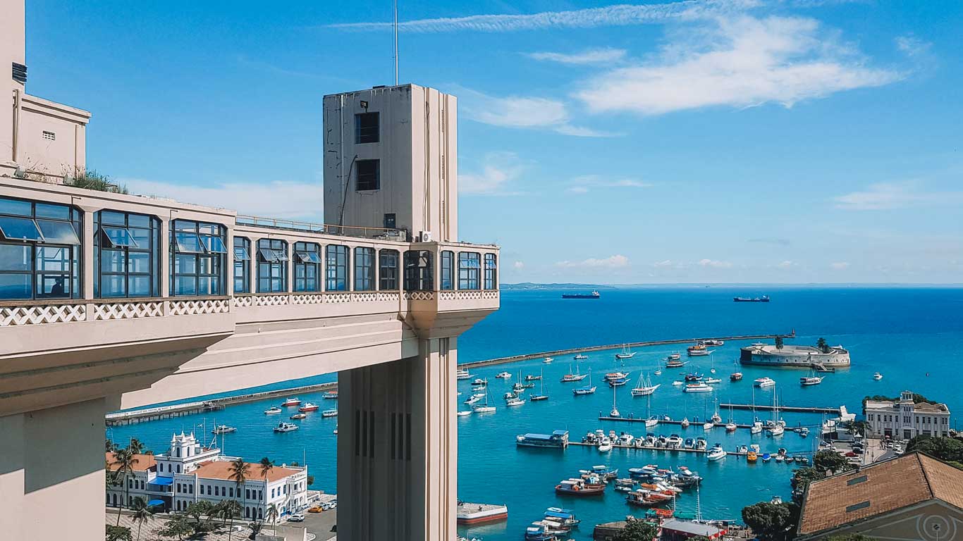 This image shows Salvador's iconic Lacerda Elevator, a striking vertical structure overlooking a vibrant bay with boats and a marina. The turquoise waters stretch to the horizon under a bright blue sky, with historic white buildings and lush greenery in the foreground.