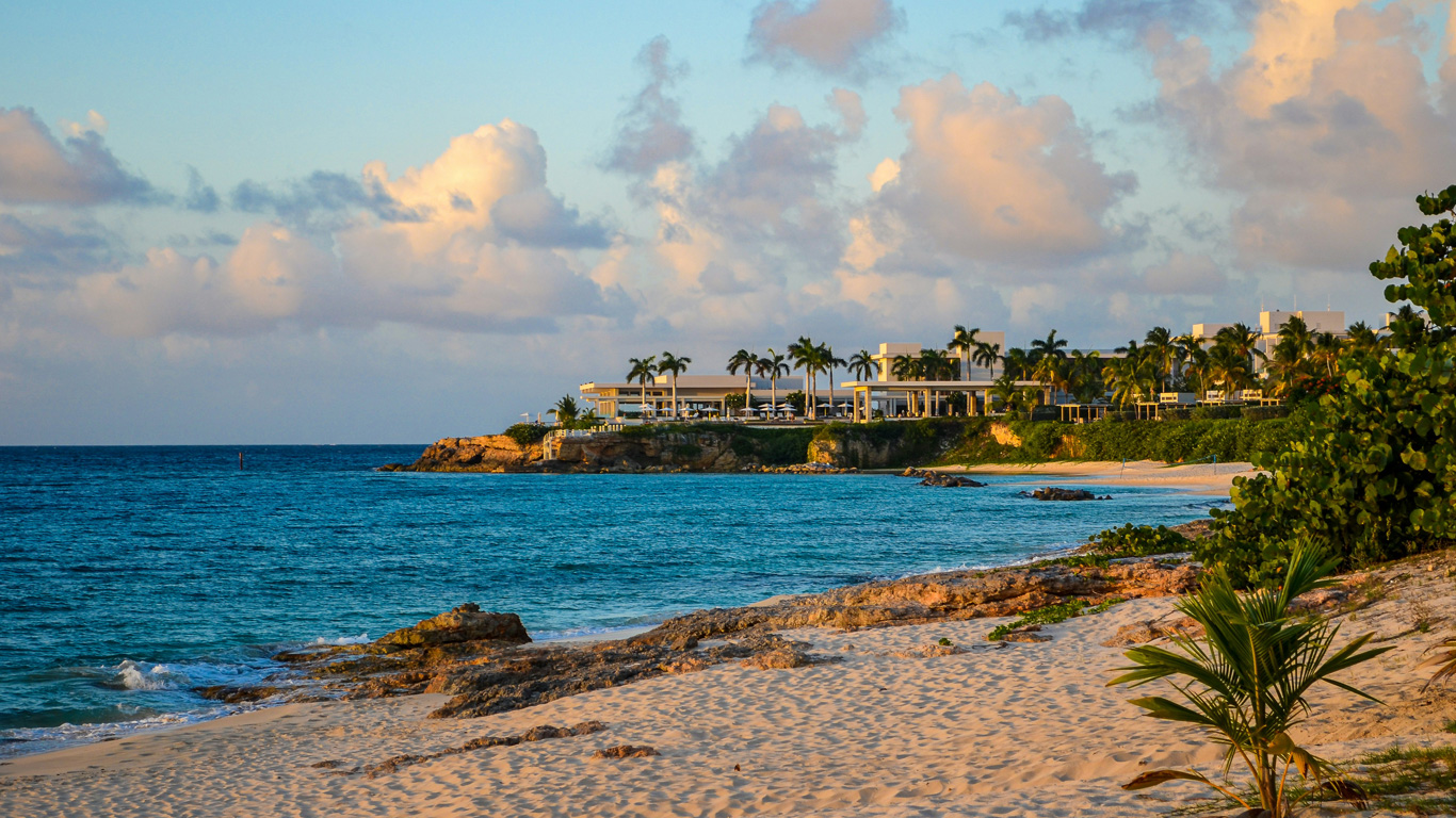 Golden sunset light casts a warm glow over a pristine beach in Anguilla, with turquoise waves gently washing onto the soft sand. A luxury beachfront resort sits atop a rocky cliff, surrounded by swaying palm trees and lush greenery. The sky is dotted with fluffy clouds, adding to the serene and tropical ambiance.