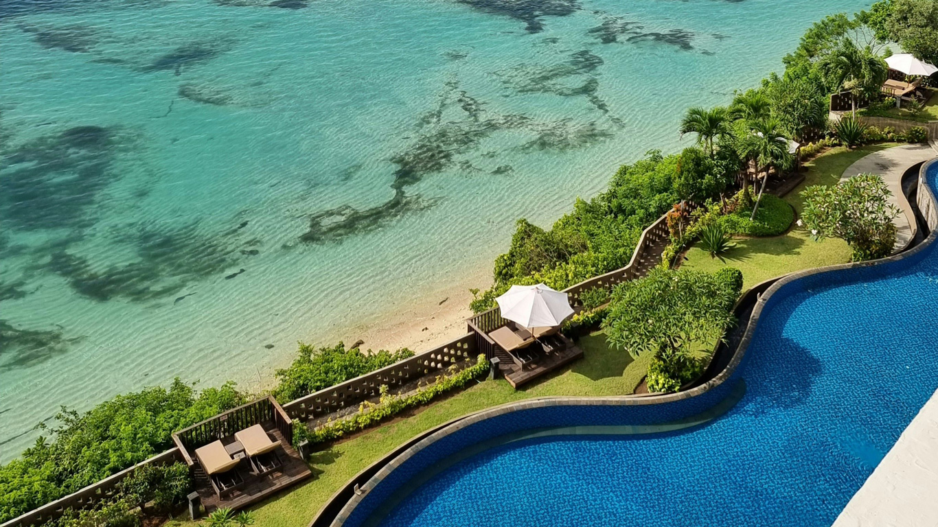 Luxury tropical resort in Bali, Indonesia, featuring an infinity pool overlooking a pristine beach with crystal-clear turquoise waters. Lush greenery and private lounge areas with umbrellas add to the serene and exclusive ambiance. The calm shoreline and beautifully landscaped surroundings create a perfect paradise retreat.