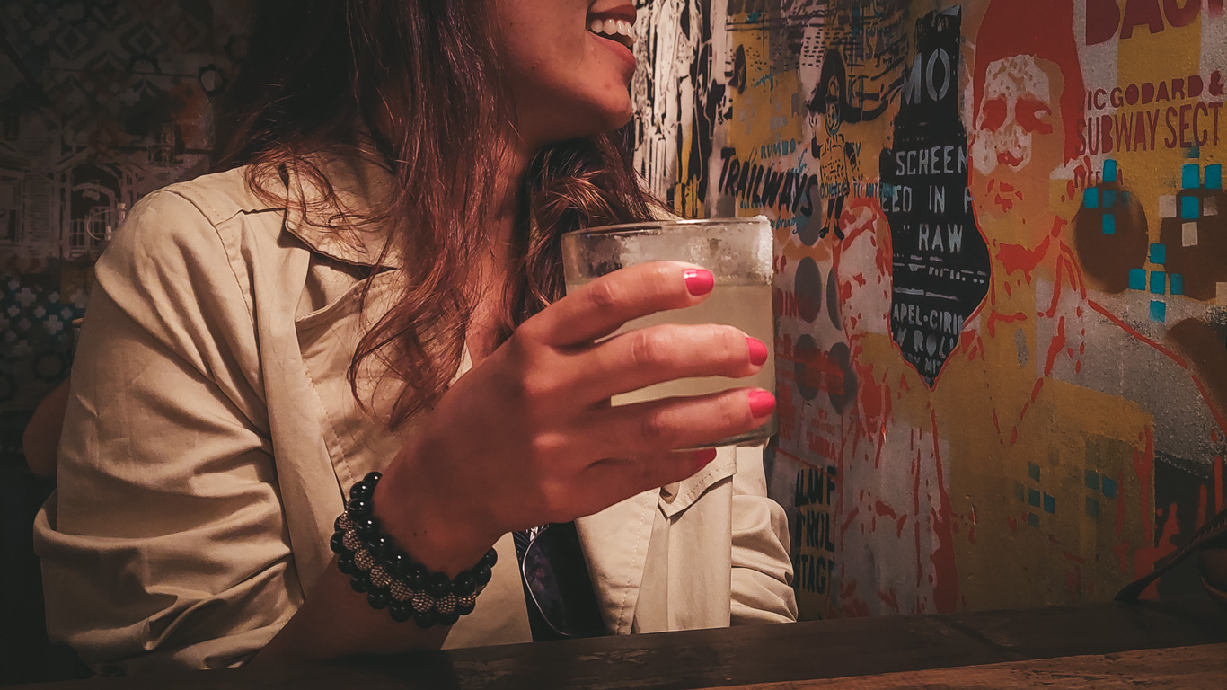 A woman with long brown hair and pink-painted nails holds a glass of a cold cocktail while smiling at a bar in Palermo, Buenos Aires. She wears a beige trench coat and a beaded black bracelet, sitting against a graffiti-covered wall with vibrant stencil art and retro-style typography. The dimly lit, artsy atmosphere suggests a trendy bar or nightclub setting.