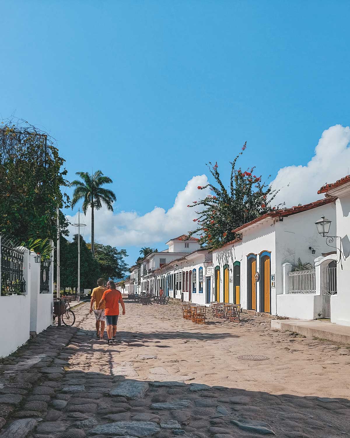 A picturesque cobblestone street in Paraty, Brazil, lined with white colonial-style buildings featuring colorful doors and windows. Two casually dressed people walk hand in hand down the street, while outdoor tables and a bicycle lean against a fence, adding to the relaxed atmosphere. The bright blue sky, palm trees, and flowering plants enhance the historic town’s charm.