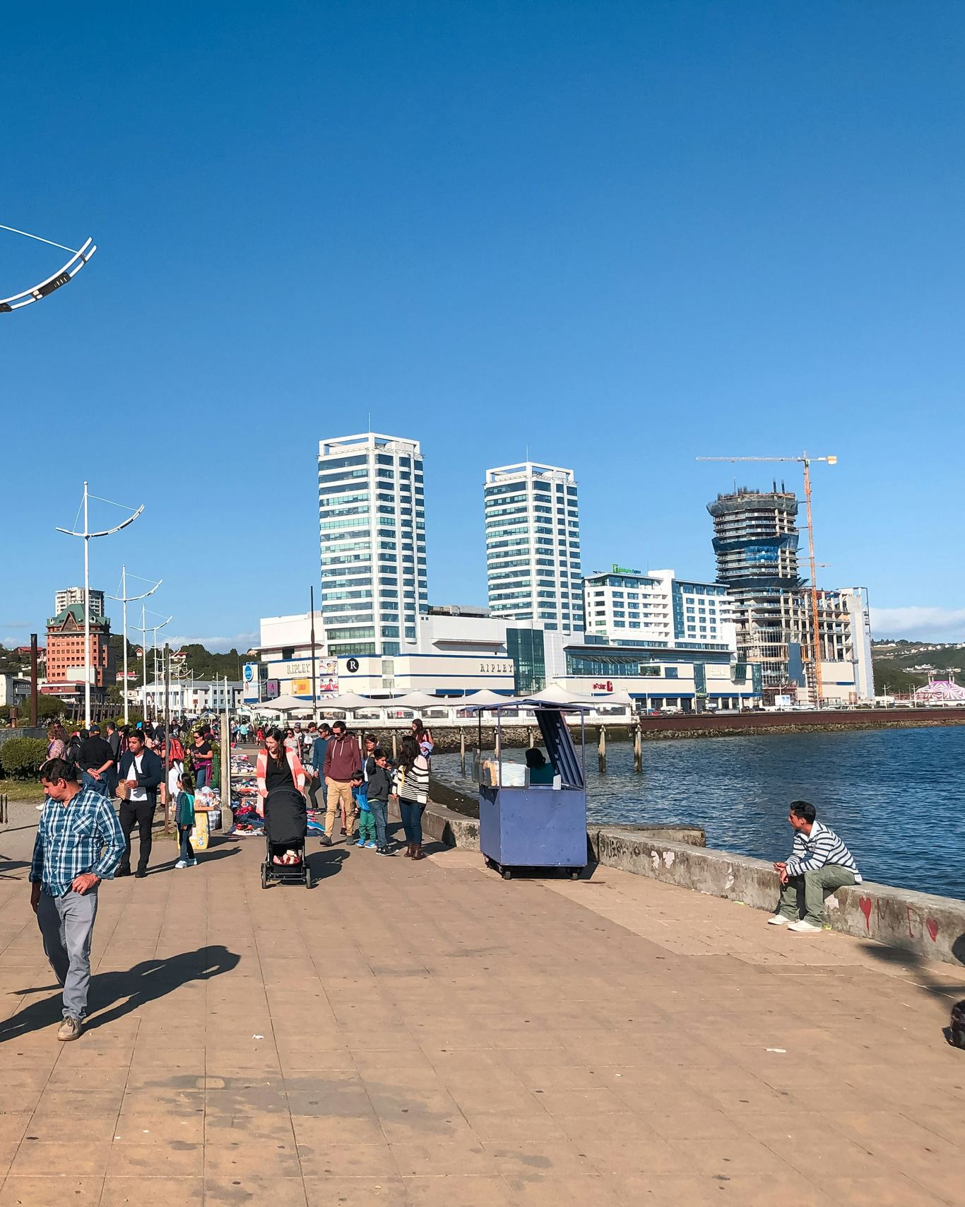 A sunny waterfront promenade in Puerto Montt, Chile, with people walking, shopping, and enjoying the view of the ocean. A small blue street cart vendor stands near the water, while a man sits on the edge of the pier. In the background, modern high-rise buildings, a shopping mall, and a construction site with a crane contrast with the lively pedestrian walkway.