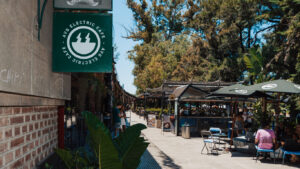 Outdoor café scene in Palermo, Buenos Aires, with a green sign reading 'AVG Electric Café' hanging from a brick and stone building. The pedestrian walkway is lined with lush greenery, shaded seating areas, and a kiosk serving coffee. People are sitting at tables under large umbrellas, chatting and enjoying the sunny day, while others walk along the vibrant open-air market space.