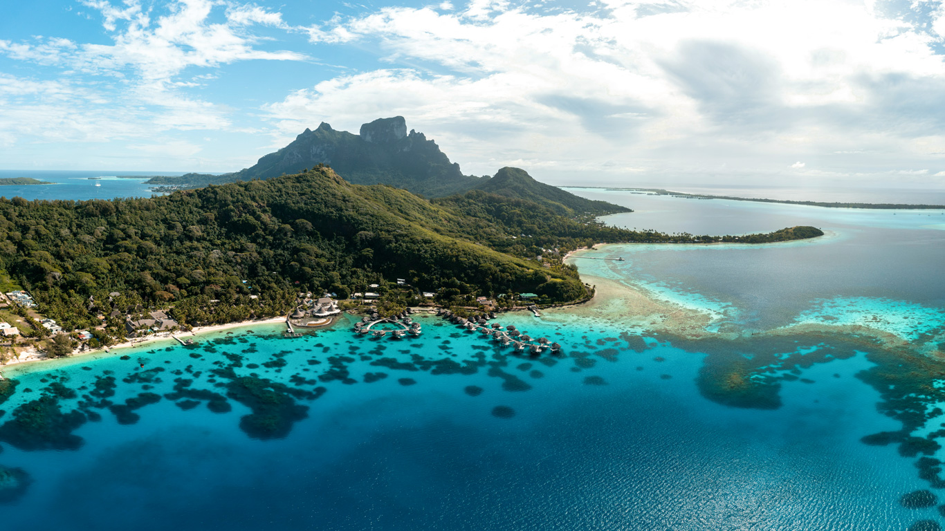 A breathtaking aerial view of Bora Bora, French Polynesia, showcasing lush green mountains, overwater bungalows, and vibrant turquoise lagoons. The crystal-clear waters reveal coral reefs beneath the surface, creating a mesmerizing contrast against the deep blue ocean. The island’s tropical landscape and luxurious resorts highlight its reputation as a dream travel destination.