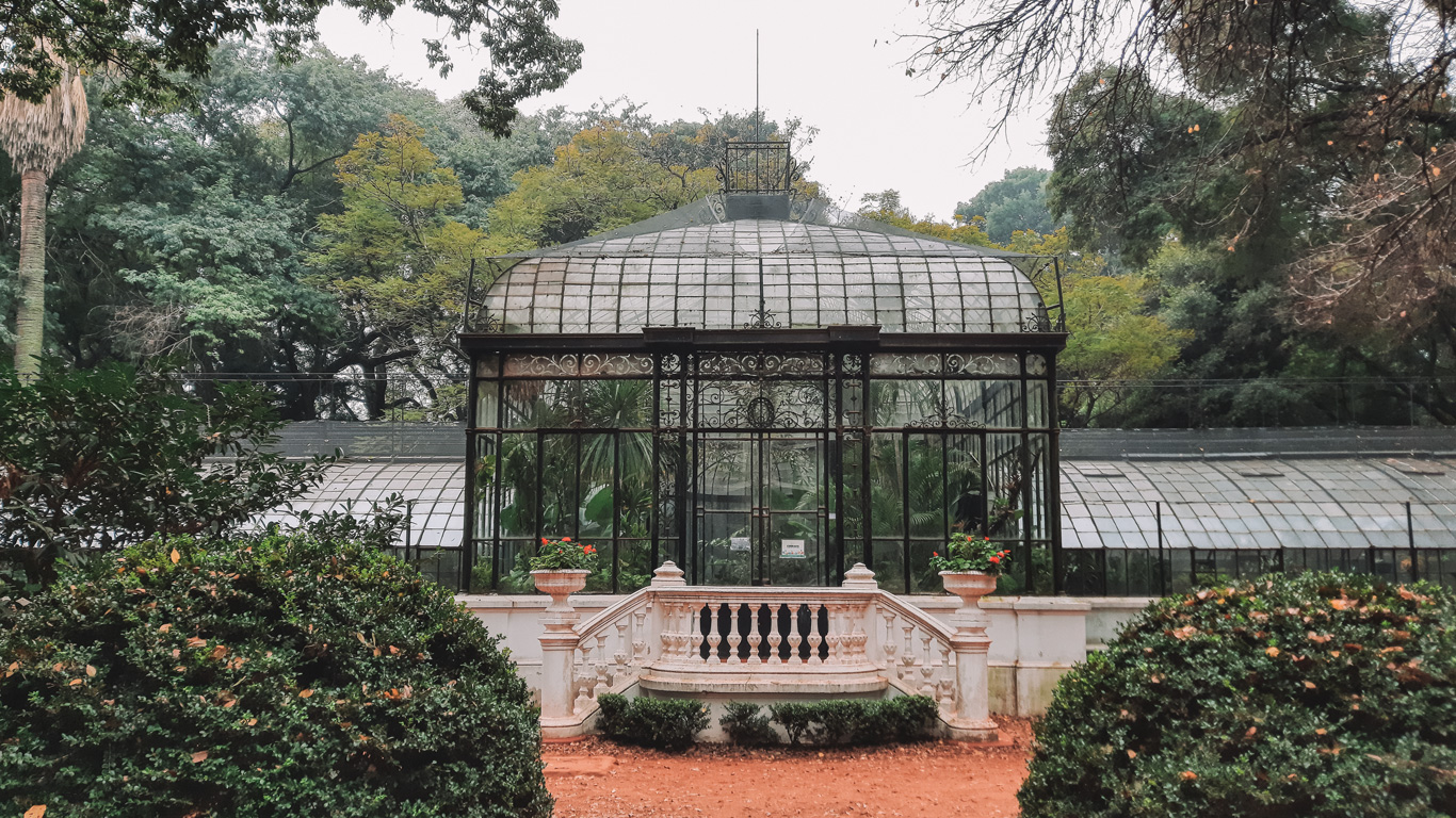 Elegant glass greenhouse with intricate black wrought iron detailing, located in the Botanical Garden of Palermo, Buenos Aires. The entrance features a white balustrade staircase adorned with red flower planters, surrounded by lush greenery and towering trees. The greenhouse’s transparent panels reveal an abundance of tropical plants inside, blending seamlessly with the verdant park setting.