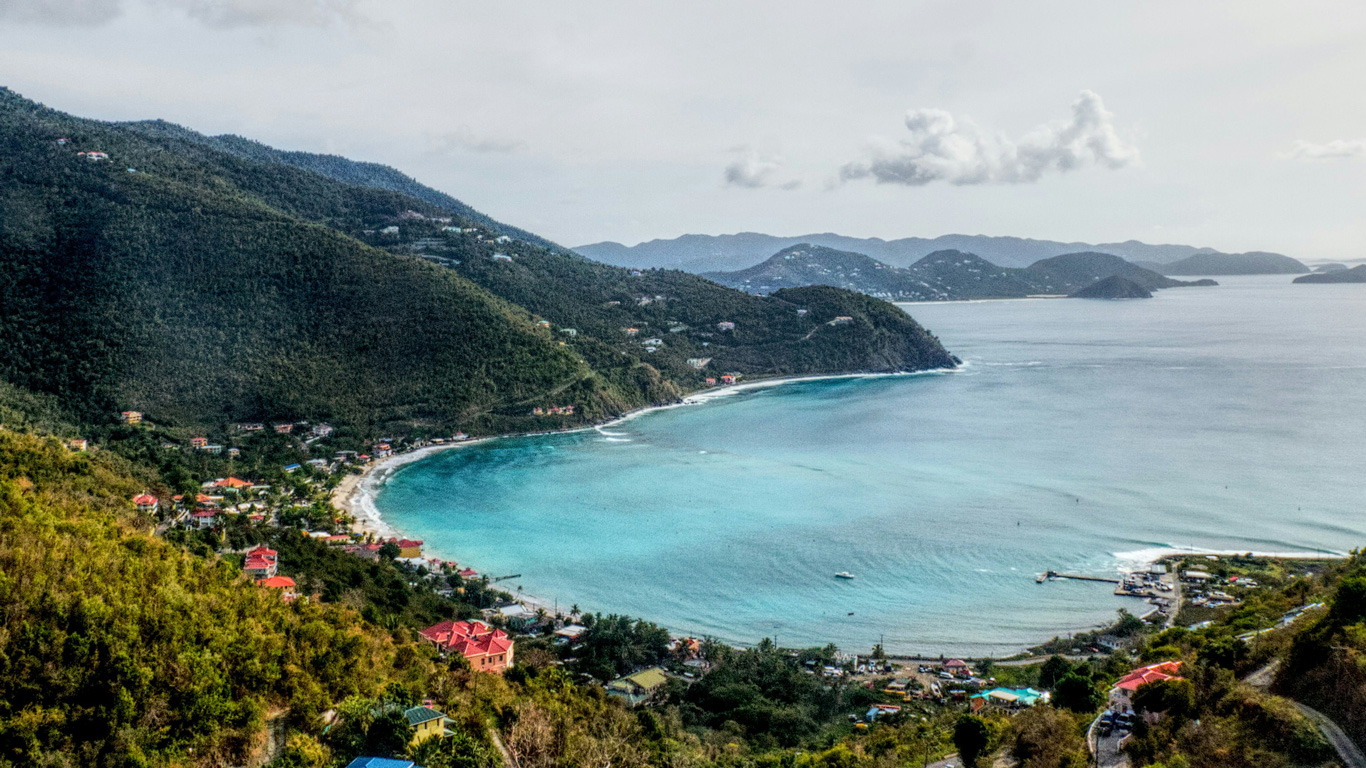 A breathtaking aerial view of a secluded bay in the British Virgin Islands, with lush green hillsides surrounding the turquoise waters. The crescent-shaped beach is dotted with colorful houses and small boats anchored near the shore. The rolling landscape and distant islands in the background add to the serene, tropical paradise setting.