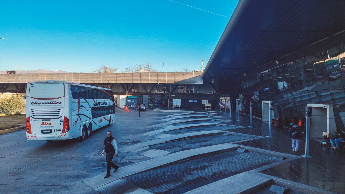 A bus terminal with a large white and red Chevallier Mix long-distance bus parked near the platform. A few passengers are waiting near numbered boarding gates, while a man in a vest walks across the pavement. The terminal’s reflective glass wall mirrors the buses and people, adding depth to the modern structure.