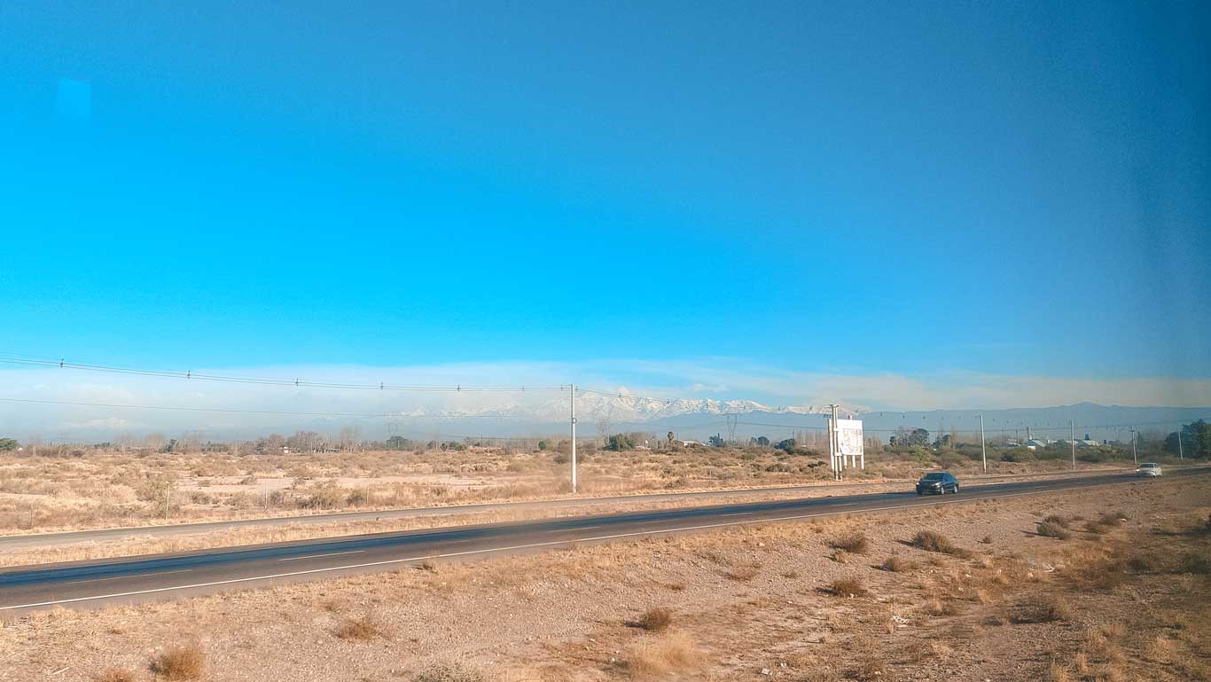 A highway cutting through a dry, arid landscape with sparse vegetation, leading toward the distant snow-capped Andes mountains. A few cars travel along the road under a clear blue sky, with power lines stretching across the horizon. The scene captures the journey through the vast Argentine countryside on the way to Mendoza.